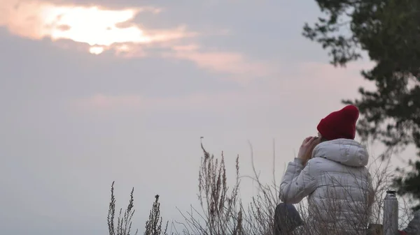 Μια ελκυστική νεαρή γυναίκα σε ένα κόκκινο καπέλο βόλτες μέσα στο δάσος, στην αρχή της άνοιξης με ένα μεγάλο τουριστικό σακίδιο, ποτά τσαγιού από ένα θερμός — Φωτογραφία Αρχείου