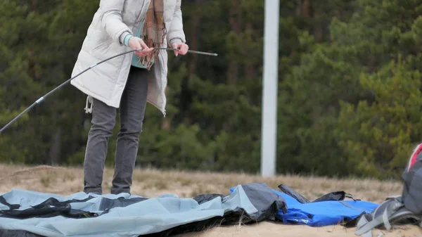 Mulher turística jovem atraente em um chapéu vermelho recolhe uma barraca turística perto da floresta na costa — Fotografia de Stock