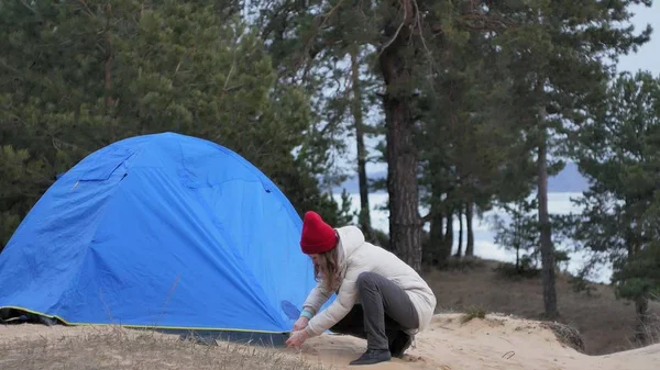Привлекательная молодая туристка в красной шляпе собирает туристическую палатку возле леса на побережье — стоковое фото