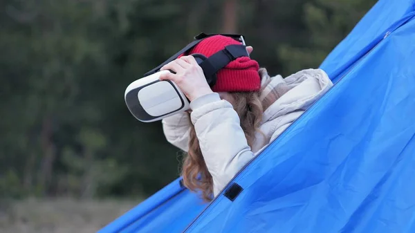 Atraente jovem turista em um chapéu vermelho senta-se em uma tenda turística e goza de um capacete realidade virtual — Fotografia de Stock