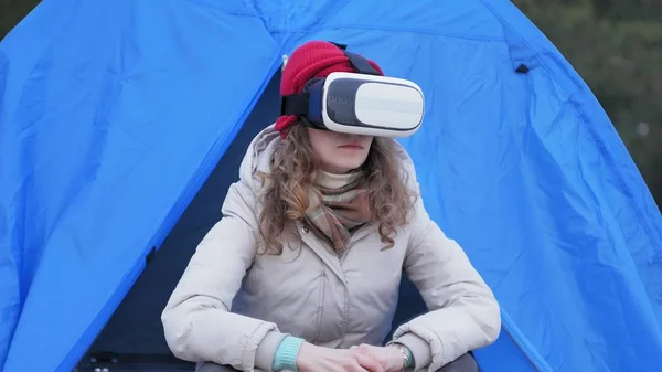 Atraente jovem turista em um chapéu vermelho senta-se em uma tenda turística e goza de um capacete realidade virtual — Fotografia de Stock