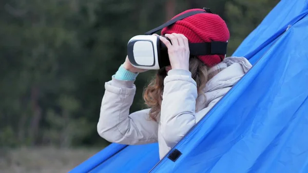 Atraente jovem turista em um chapéu vermelho senta-se em uma tenda turística e goza de um capacete realidade virtual — Fotografia de Stock