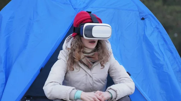 Atraente jovem turista em um chapéu vermelho senta-se em uma tenda turística e goza de um capacete realidade virtual — Fotografia de Stock