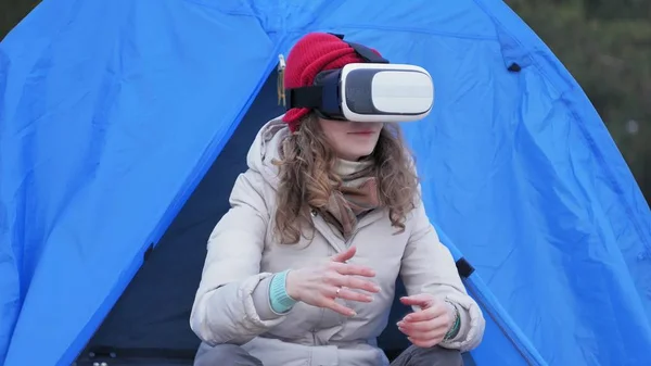 Atraente jovem turista em um chapéu vermelho senta-se em uma tenda turística e goza de um capacete realidade virtual — Fotografia de Stock