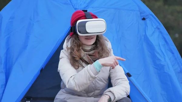 Atraente jovem turista em um chapéu vermelho senta-se em uma tenda turística e goza de um capacete realidade virtual — Fotografia de Stock