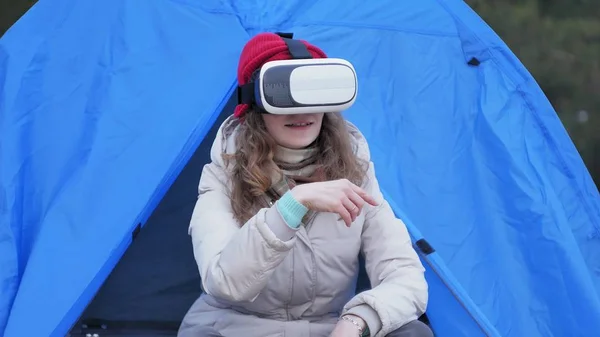 Atraente jovem turista em um chapéu vermelho senta-se em uma tenda turística e goza de um capacete realidade virtual — Fotografia de Stock