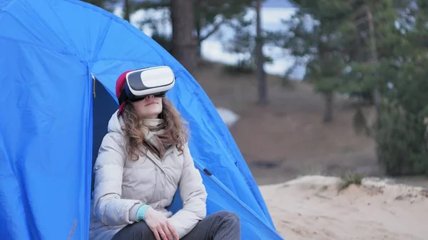 Atraente jovem turista em um chapéu vermelho senta-se em uma tenda turística e goza de um capacete realidade virtual — Fotografia de Stock