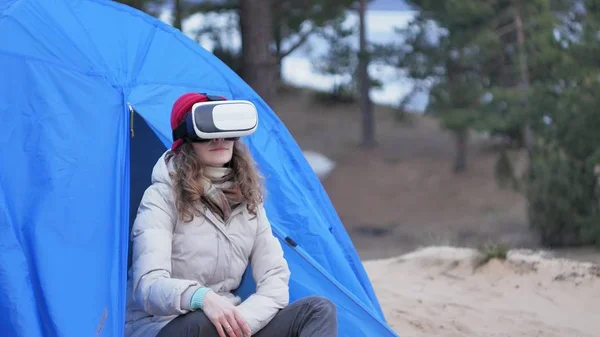 Atraente jovem turista em um chapéu vermelho senta-se em uma tenda turística e goza de um capacete realidade virtual — Fotografia de Stock