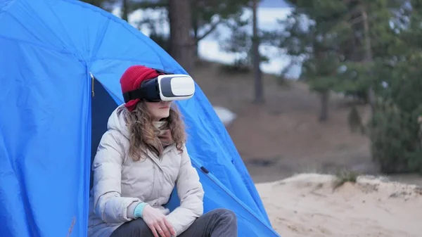 Atraente jovem turista em um chapéu vermelho senta-se em uma tenda turística e goza de um capacete realidade virtual — Fotografia de Stock