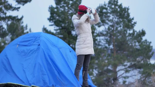 Attraente giovane donna turista con un cappello rosso si siede in una tenda turistica e gode di un casco realtà virtuale — Foto Stock