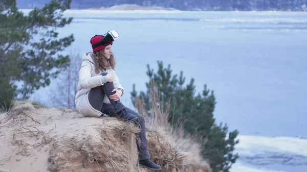 Atractiva joven turista en un sombrero rojo se sienta en una en la orilla y disfruta de un casco de realidad virtual — Foto de Stock