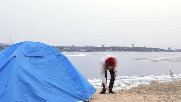 Attraente giovane donna turistica con un cappello rosso raccoglie una tenda turistica vicino alla foresta sulla costa — Foto Stock