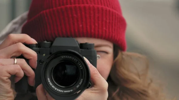 Νεαρή όμορφη γυναίκα σε ένα κόκκινο καπέλο που φοράει σπορ ζεστά ρούχα και οδοστρωτήρες, κάθεται σε ένα ξύλινο πάγκο και λήψη φωτογραφιών σε μια vintage φωτογραφική μηχανή — Φωτογραφία Αρχείου