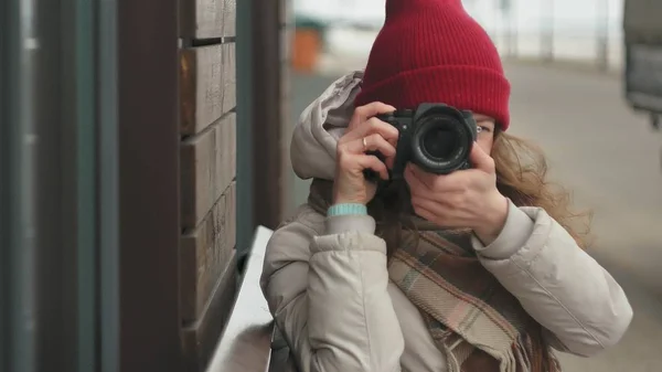 Young beautiful woman in a red hat wearing sporty warm clothes and rollers, sitting on a wooden bench and taking pictures on a vintage camera — Stock Photo, Image
