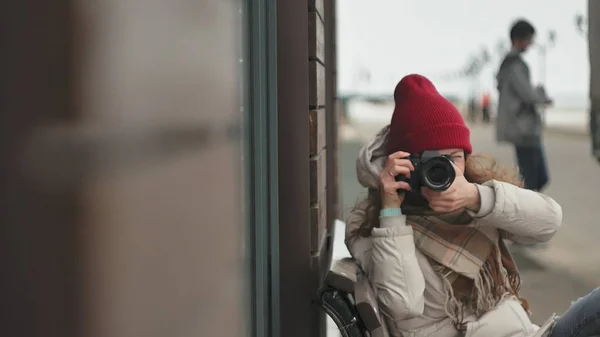 Mooie jongedame in een rode hoed dragen sportieve warme kleren en rollen, zittend op een houten bankje en nemen van foto's op een vintage camera — Stockfoto