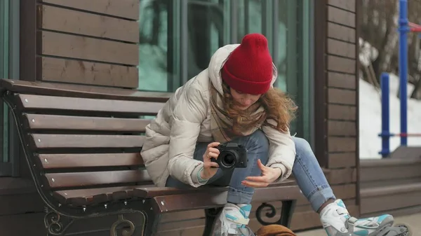 Jovem mulher bonita em um chapéu vermelho vestindo roupas e rolos quentes esportivos, sentado em um banco de madeira e tirando fotos em uma câmera vintage — Fotografia de Stock
