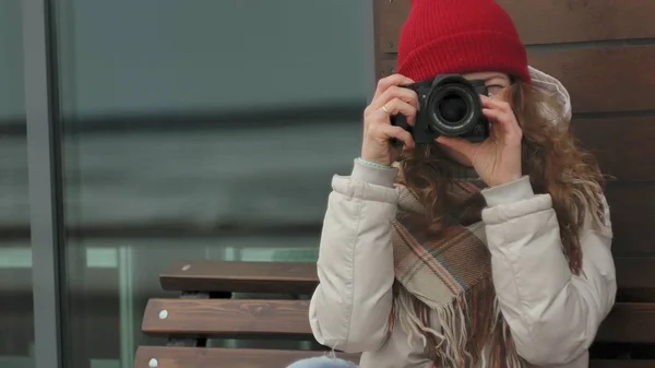 Young beautiful woman in a red hat wearing sporty warm clothes and rollers, sitting on a wooden bench and taking pictures on a vintage camera — Stock Photo, Image