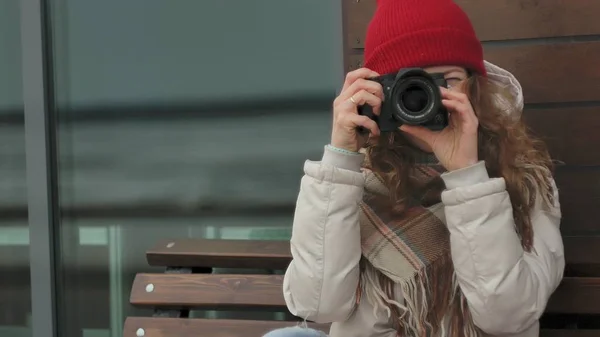 Mooie jongedame in een rode hoed dragen sportieve warme kleren en rollen, zittend op een houten bankje en nemen van foto's op een vintage camera — Stockfoto