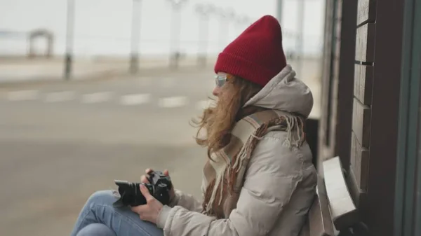 Mooie jongedame in een rode hoed dragen sportieve warme kleren en rollen, zittend op een houten bankje en nemen van foto's op een vintage camera — Stockfoto