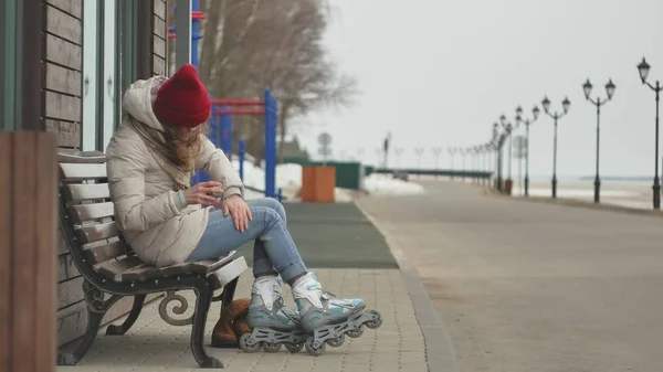 Giovane bella donna con un cappello rosso che indossa vestiti caldi sportivi e rulli, seduta su una panchina di legno bere il tè da un thermos — Foto Stock