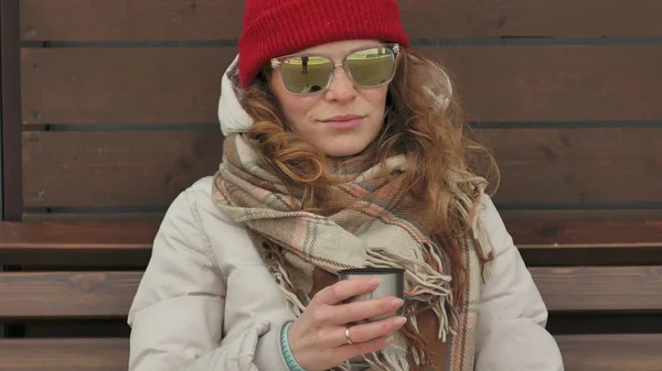 Νεαρή όμορφη γυναίκα σε ένα κόκκινο καπέλο που φοράει σπορ ζεστά ρούχα και οδοστρωτήρες, κάθεται σε ένα ξύλινο παγκάκι πίνοντας τσάι από ένα θερμός — Φωτογραφία Αρχείου