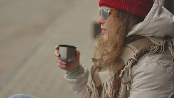 Young beautiful woman in a red hat wearing sporty warm clothes and rollers, sitting on a wooden bench drinking tea from a thermos — Stock Photo, Image