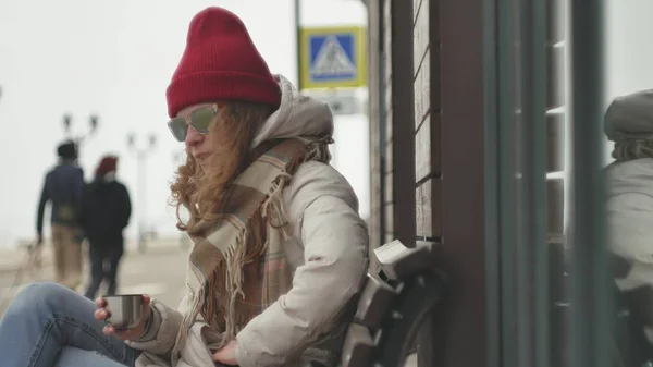 Young beautiful woman in a red hat wearing sporty warm clothes and rollers, sitting on a wooden bench drinking tea from a thermos — Stock Photo, Image