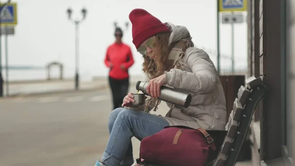 Junge schöne Frau mit rotem Hut in sportlich warmer Kleidung und Rollschuhen, die auf einer Holzbank sitzt und Tee aus einer Thermoskanne trinkt — Stockfoto