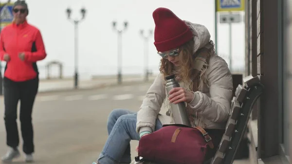 Mooie jongedame in een rode hoed dragen sportieve warme kleren en rollen, zittend op een houten bankje thee drinken uit een thermos — Stockfoto