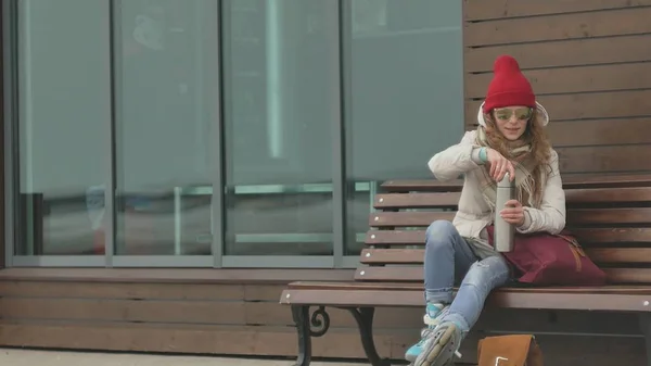 Young beautiful woman in a red hat wearing sporty warm clothes and rollers, sitting on a wooden bench drinking tea from a thermos — Stock Photo, Image