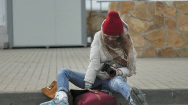 Kırmızı şapka giyen sportif sıcak giysiler ve silindirleri, asfalt yolda ve vintage fotoğraf makinelerinde fotoğraf çekimi oturan genç güzel kadın