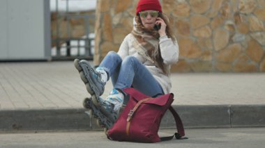 Kırmızı bir şapka giyen sportif sıcak giysiler ve Silindirler, asfalt yol üzerinde oturan ve telefonda konuşurken genç güzel kadın