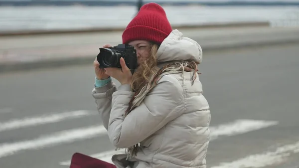 Junge schöne Frau mit rotem Hut in sportlich warmer Kleidung und Rollschuhen, die auf der Asphaltstraße sitzt und mit einer Oldtimer-Kamera Fotos macht — Stockfoto