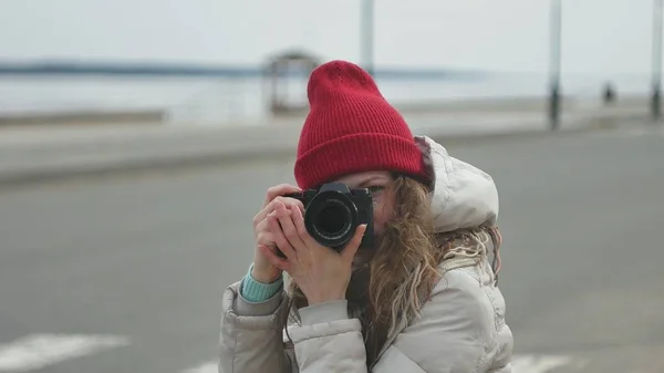 Junge schöne Frau mit rotem Hut in sportlich warmer Kleidung und Rollschuhen, die auf der Asphaltstraße sitzt und mit einer Oldtimer-Kamera Fotos macht — Stockfoto