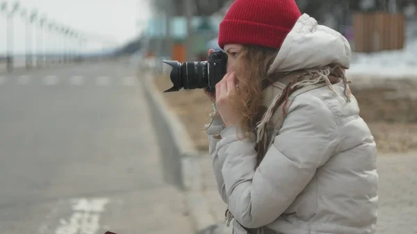 Junge schöne Frau mit rotem Hut in sportlich warmer Kleidung und Rollschuhen, die auf der Asphaltstraße sitzt und mit einer Oldtimer-Kamera Fotos macht — Stockfoto