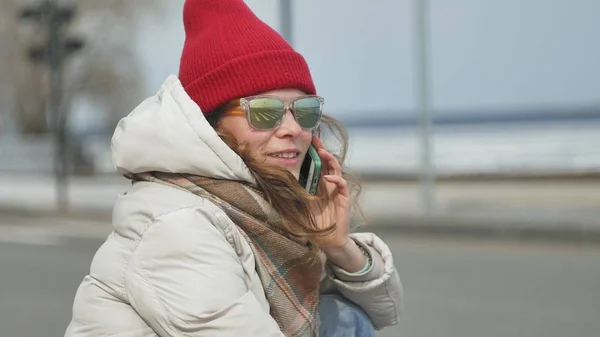 Ung vacker kvinna i en röd hatt bär sportig varma kläder och rullar, sitter på asfaltvägen och prata i telefon — Stockfoto