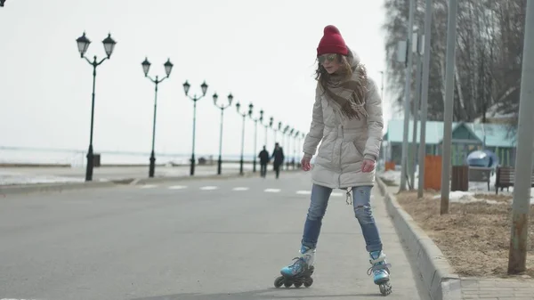 Fiatal gyönyörű nő egy piros sapkát visel, sportos meleg ruhát és a görgők, lovaglás a tengerparton úton — Stock Fotó