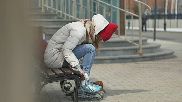 Young beautiful woman in sporty warm clothes and rollers, sitting on a wooden bench and dresses roller skates getting ready with skating — Stock Photo, Image
