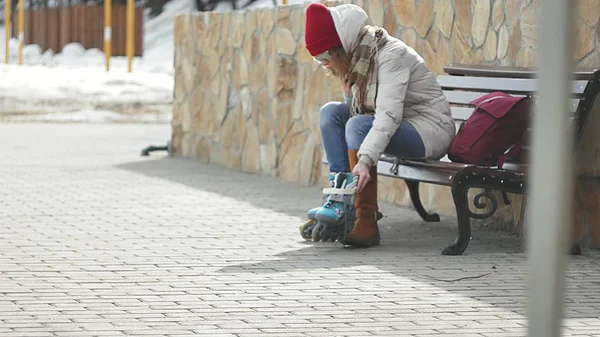 Joven mujer hermosa en ropa de abrigo deportivo y rodillos, sentado en un banco de madera y vestidos patines preparándose con patinaje — Foto de Stock