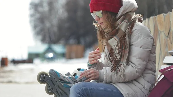 Young beautiful woman in sporty warm clothes and rollers, sitting on a wooden bench and dresses roller skates getting ready with skating — Stock Photo, Image