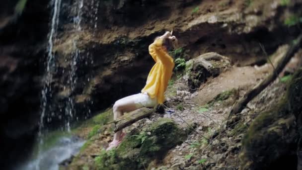 Young woman standing in front of waterfall with her hands raised. Female tourist with her arms outstretched looking at waterfall. — Stock Video