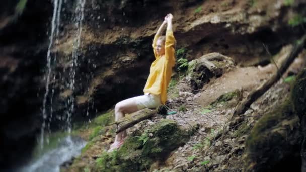 Mujer joven de pie frente a la cascada con las manos levantadas. Turista femenina con los brazos extendidos mirando la cascada . — Vídeos de Stock