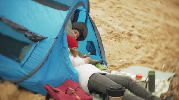 Turist-tältet på flodstranden. två flicka sitter nära tältet ätande och drickande ser vid floden. — Stockvideo