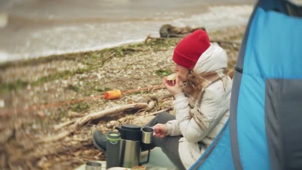 Toeristische tent aan de rivieroever. twee meisje zit in de buurt van de tent, eten en drinken kijkt naar de rivier. — Stockvideo