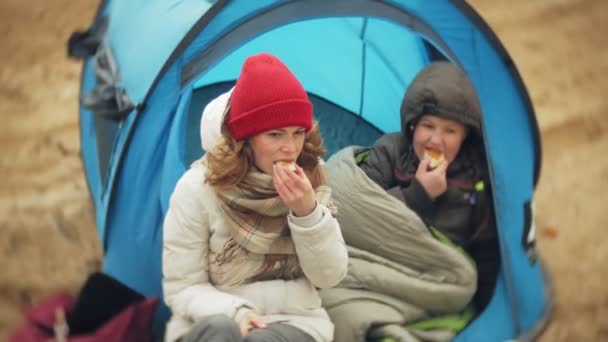 Туристическая палатка на берегу реки. две девочки сидят возле палатки, едят и пьют, смотрят на реку . — стоковое видео