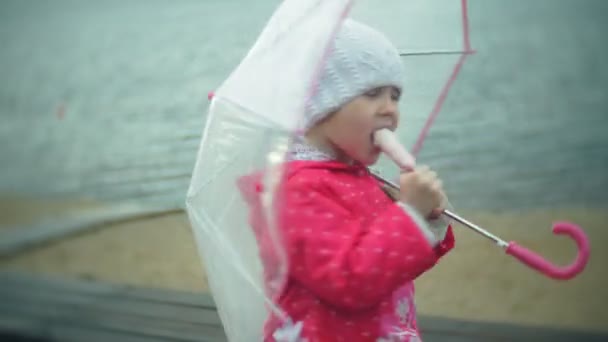 Pequena menina bonita com guarda-chuva jogando na chuva comendo sorvete na costa — Vídeo de Stock