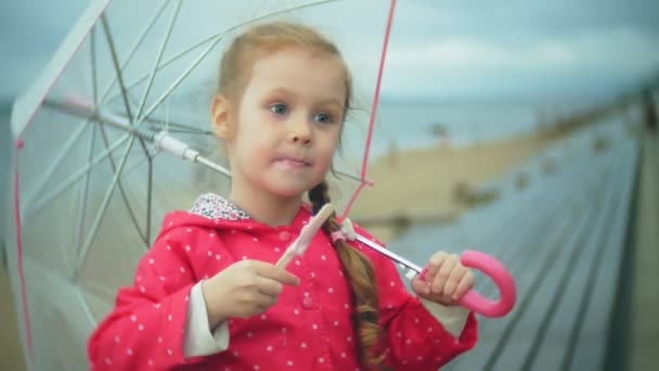 Niña hermosa con paraguas jugando bajo la lluvia comiendo helado en la costa — Vídeos de Stock