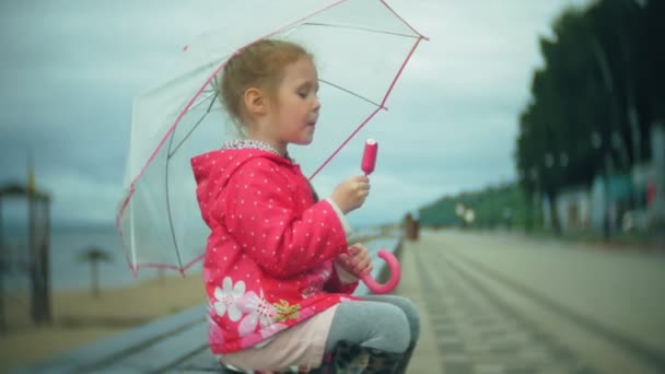 Niña hermosa con paraguas jugando bajo la lluvia comiendo helado en la costa — Vídeos de Stock