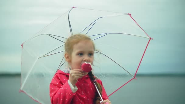Pequena menina bonita com guarda-chuva jogando na chuva comendo sorvete na costa — Vídeo de Stock