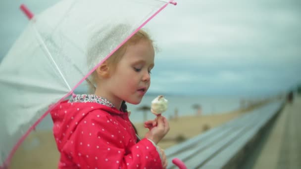 Piccola bella ragazza con ombrello che gioca sotto la pioggia mangiando gelato sulla costa — Video Stock